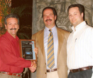 three men in suites, two shaking hands, accepting an award